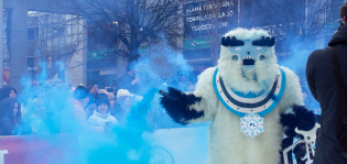 Jeti-maskotti Pikku on laskenut pulkkakisan ja pitelee dramaattisesti savuavaa savutehostetta kädessään.